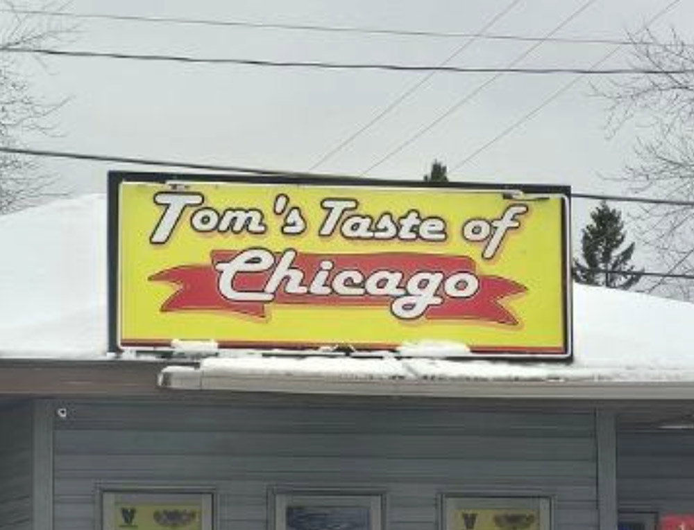 A yellow and red sign on a building reads “Tom's Taste of Chicago.” The roof below the sign is covered with snow, and there are leafless trees and power lines in the background.
