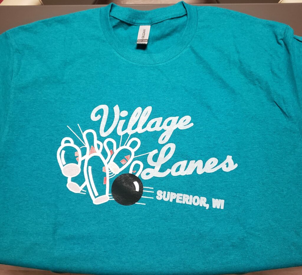 Teal T-shirt with "Village Lanes" and "Superior, WI" written in white, featuring an illustration of bowling pins being struck by a black bowling ball.