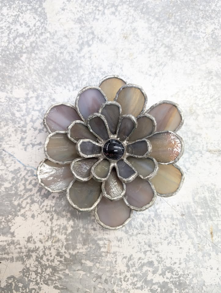 A decorative flower made of stained glass petals in shades of gray and beige, with a glossy dark center, set against a textured, light-colored background.
