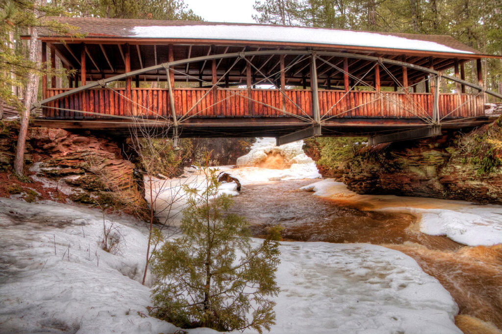 A rustic wooden covered bridge spans a partially frozen river, surrounded by snow, pine trees, and hints of spring waterfalls in the distance. The water flows under the bridge as patches of ice and snow linger on the riverbanks.