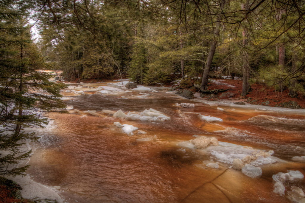 A shallow river with reddish-brown water flows through a forest of evergreens. Patches of snow and ice line the banks while distant spring waterfalls hint at the season’s early thaw.