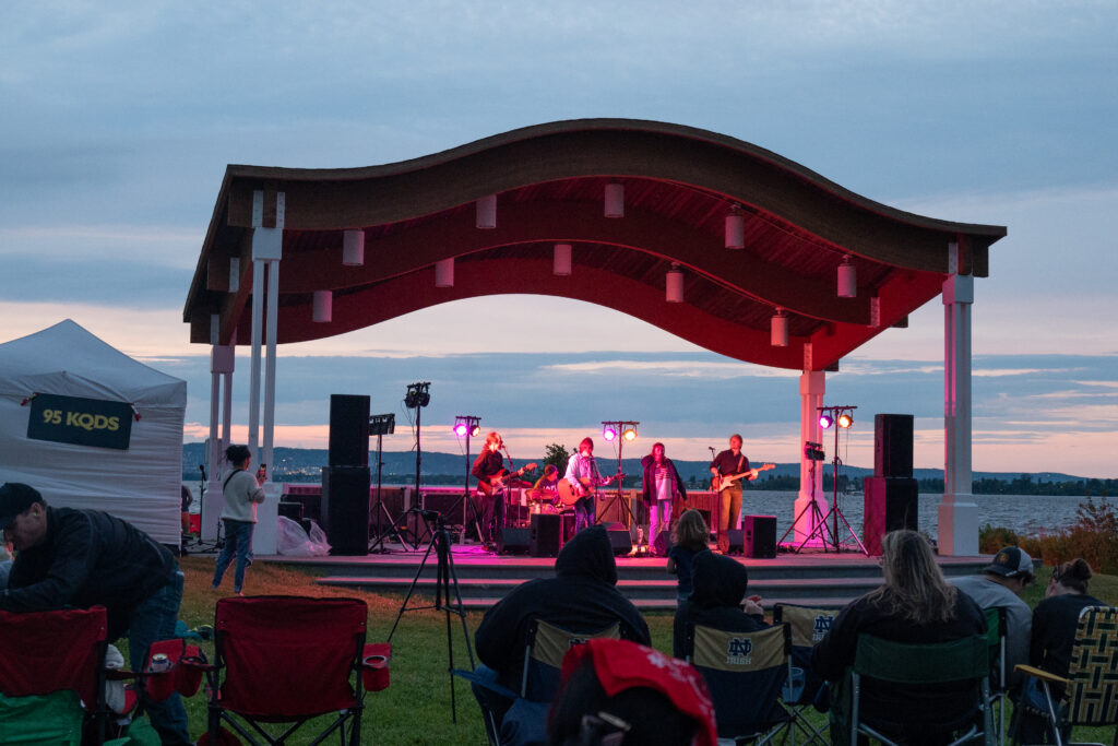 A band performs on an outdoor stage with a wavy roof at sunset. People sit in lawn chairs facing the stage, while a white tent stands to the left and spring waterfalls glitter beside the water in the background.