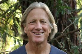 A middle-aged person with shoulder-length blond hair is smiling outdoors, standing in front of green trees and foliage.