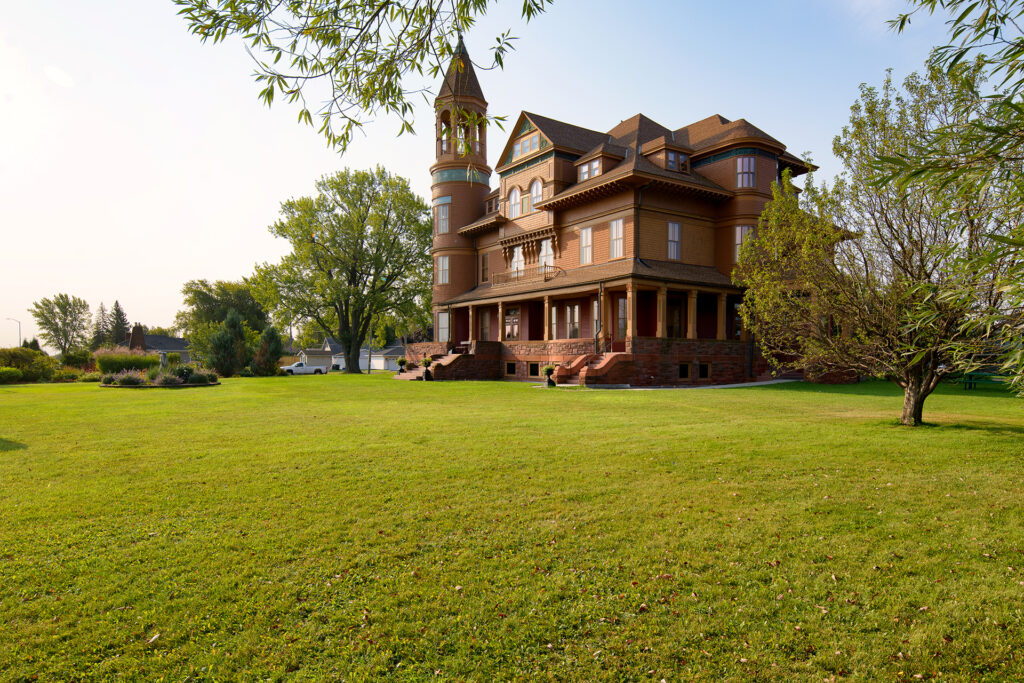 A large, historic Victorian-style house with a turret, wraparound porch, and brown exterior stands on a spacious, manicured green lawn surrounded by trees under a clear sky, evoking the fresh beauty of nearby spring waterfalls.