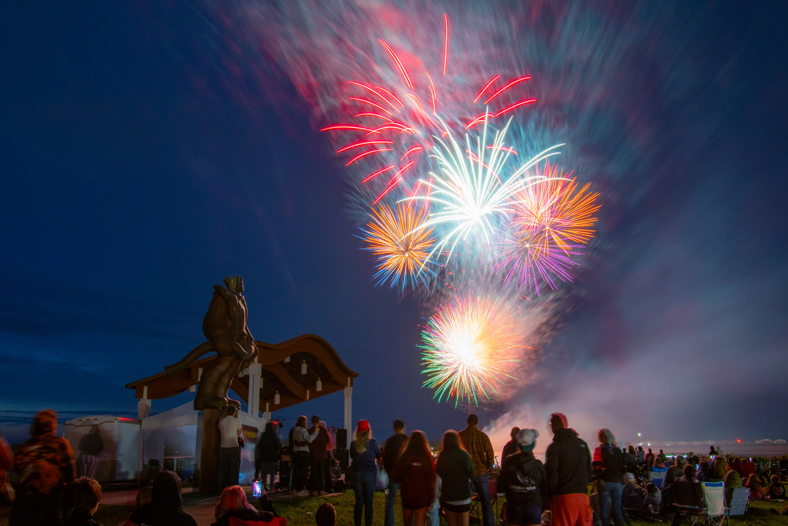 A colorful fireworks display lights up the sky over the lake and a crowd of onlookers.
