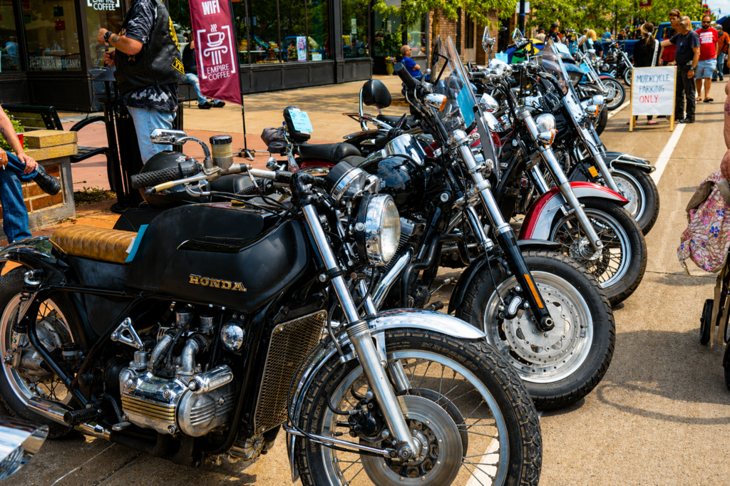 A row of parked motorcycles, including a black Honda, lines a sidewalk at an outdoor event. People walk nearby, with a sign reading "Motorcycle Parking Only" and hints of spring waterfalls in the distant background.