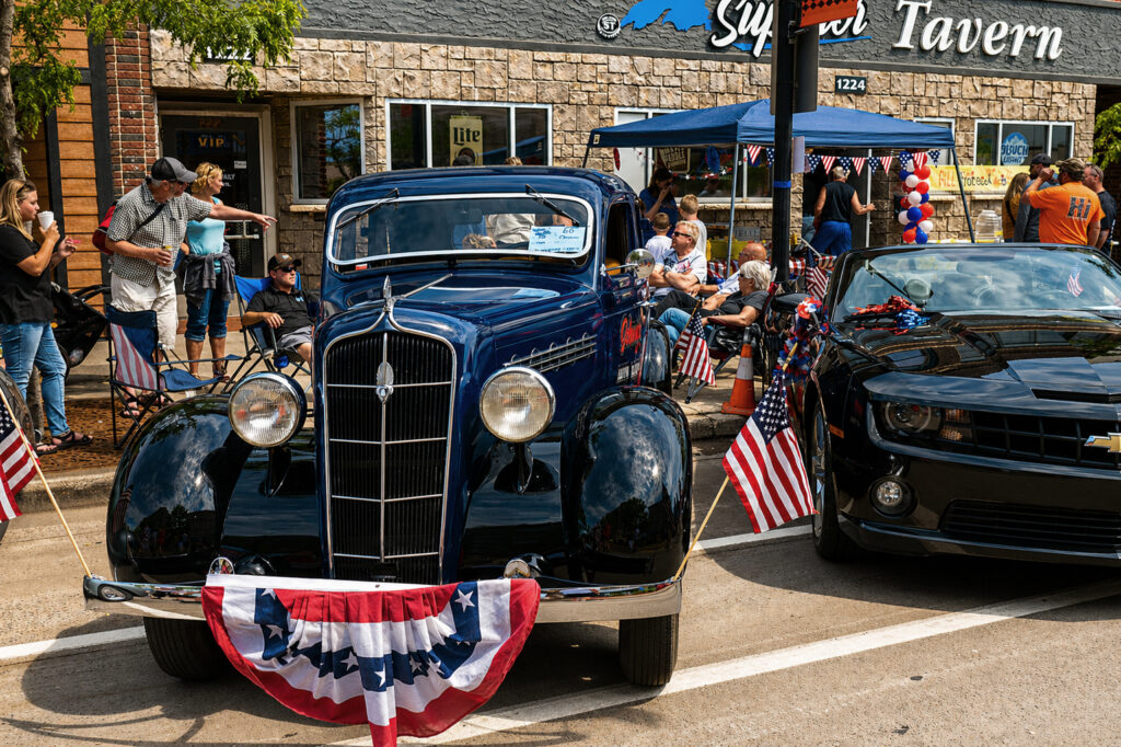 A vintage blue car adorned with American flags is parked beside a modern black car. People gather near spring waterfalls of patriotic decorations that fill the lively scene in front of a tavern.