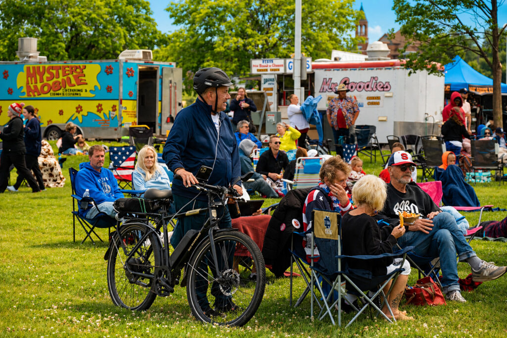A man with a helmet stands beside his bicycle on grass near a group of people sitting in chairs, as food trucks and trees set the scene for an outdoor event that feels as refreshing as spring waterfalls.