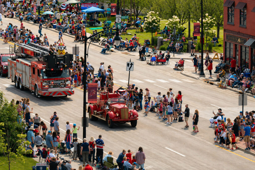 A lively parade features vintage and modern fire trucks driving down a street lined with spectators, many seated or standing, on a sunny day reminiscent of spring waterfalls. People watch from sidewalks and grassy areas, capturing the festive atmosphere.
