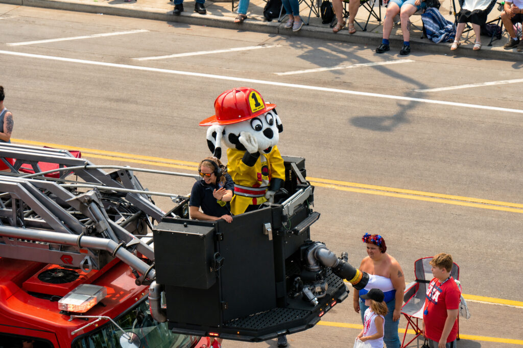 A person in a Dalmatian dog mascot costume dressed as a firefighter rides in a fire truck's aerial lift during a parade, with crowds gathered along the sidewalk under clear skies reminiscent of spring waterfalls.