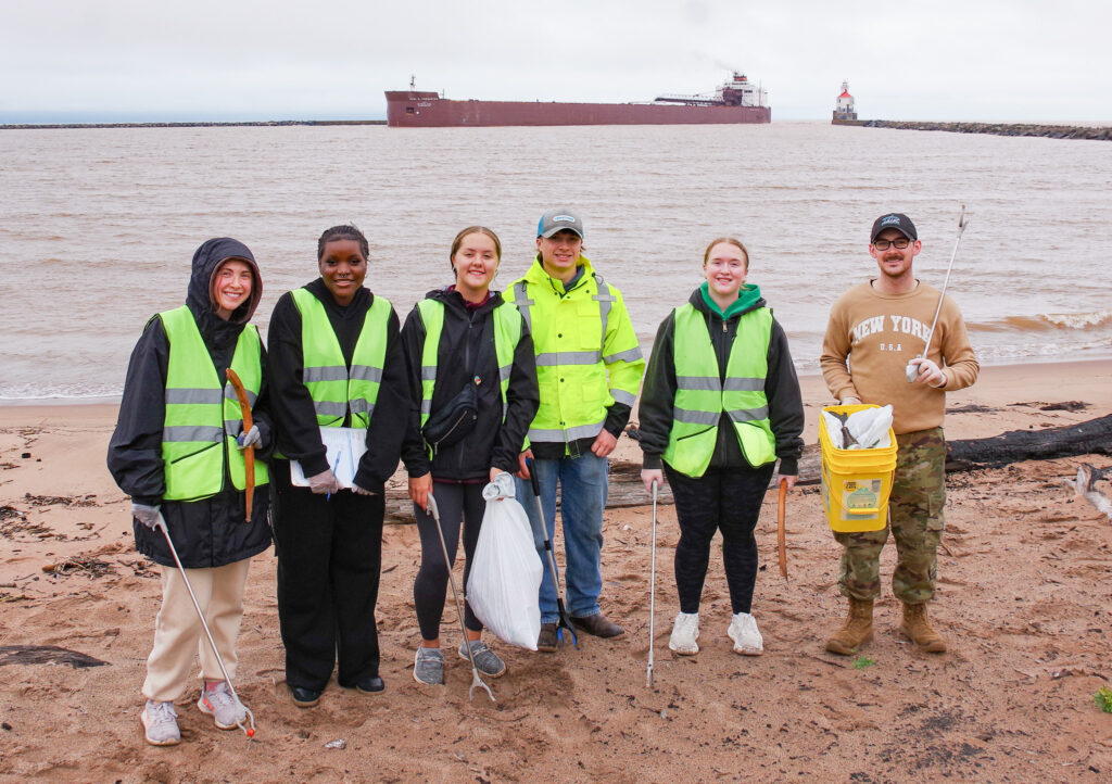 Six people wearing reflective vests and jackets stand on a sandy beach holding trash pickers and bags, participating in a cleanup. A large cargo ship is visible on the water in the background.