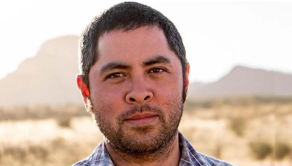 A man with short dark hair and a beard stands outdoors in a sunlit desert landscape, wearing a plaid shirt. Distant mountains are blurred in the background.