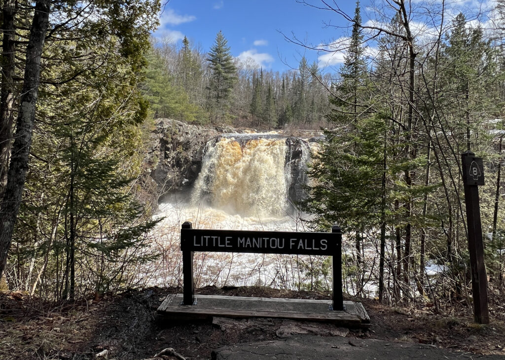 A waterfall cascades behind a sign reading "Little Manitou Falls," framed by evergreen trees under a blue sky with scattered clouds. A forest trail and trail marker on the right invite visitors to enjoy the beauty of spring waterfalls.