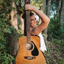 A smiling woman stands outdoors in a forested area, holding an acoustic guitar over her shoulder and looking at the camera. Sunlight filters through the trees around her.