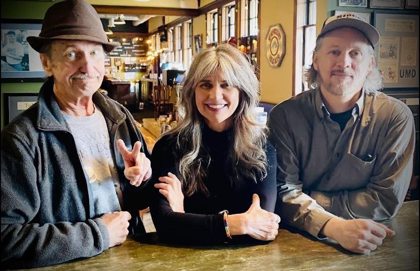 Three adults—two men and one woman—stand together at a wooden counter in a cozy, well-lit restaurant. The man on the left flashes a peace sign, while all three smile at the camera.