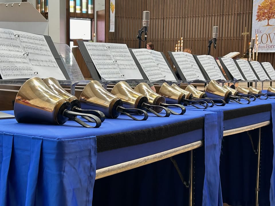 A row of handbells rests on a blue-covered table, with open sheet music stands behind them inside a church. Stained glass windows and a wooden wall are visible in the background.