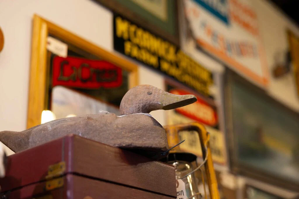 A carved wooden duck decoy sits atop a box, with various framed signs and artwork blurred in the background—a charming find while shopping in Superior, Wisconsin.