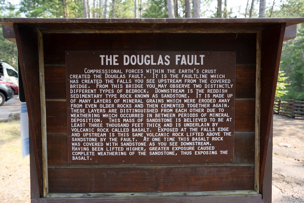 A wooden sign in a forest labeled "THE DOUGLAS FAULT" explains how compressional forces created the fault, exposing red and gray rock layers, sandstone, basalt, and nearby spring waterfalls for visitors to observe.
