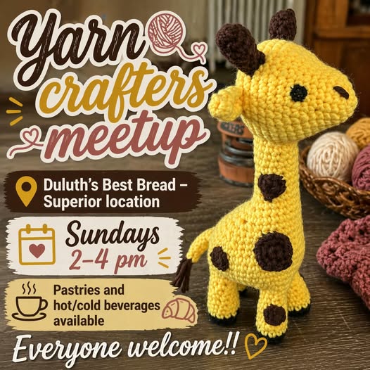 A crocheted yellow giraffe doll sits on a table surrounded by yarn and knitted items. A sign promotes a "Yarn crafters meetup" on Sundays, 2–4 pm, at Duluth’s Best Bread, with pastries and drinks available.