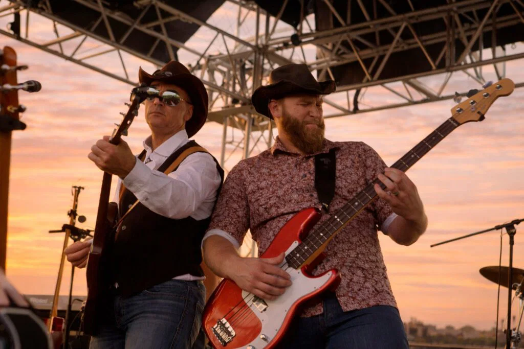 Two male musicians in cowboy hats play guitar and bass on an outdoor stage at sunset, metal scaffolding above and an orange sky behind them—capturing the lively spirit of weekend getaways in Wisconsin. One sings into a microphone while the other focuses on playing.