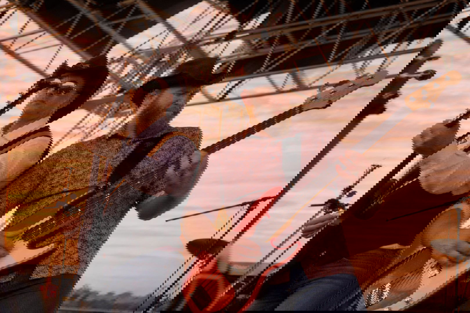 Two male musicians in cowboy hats play guitar and bass on an outdoor stage at sunset, metal scaffolding above and an orange sky behind them—capturing the lively spirit of weekend getaways in Wisconsin. One sings into a microphone while the other focuses on playing.