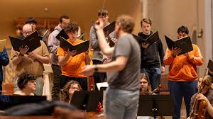 A conductor leads a choir rehearsal; several singers hold open sheet music while singing, and musicians sit in front, some playing instruments. The group is indoors, focused on their performance.