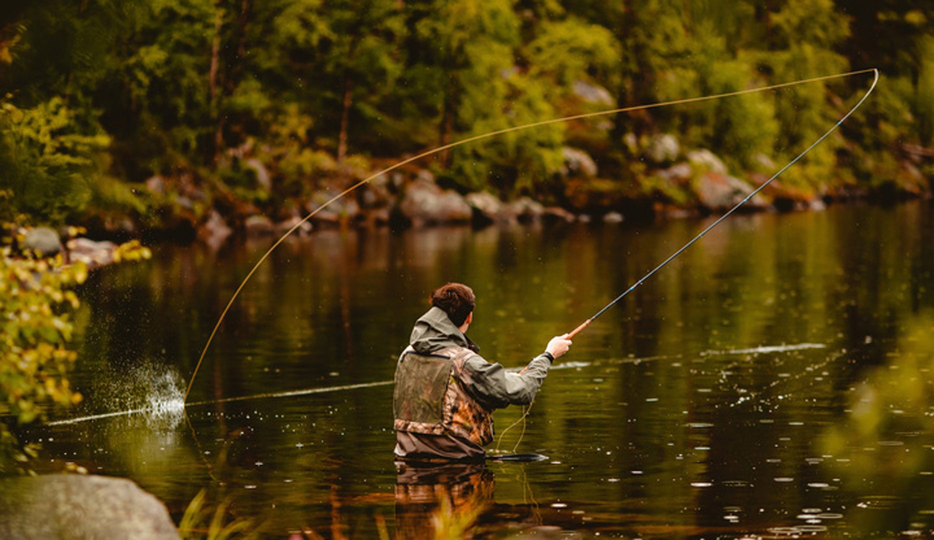 A person wearing waders fly fishes in a calm, forested river—one of many serene scenes you might find on weekend getaways in Wisconsin. Trees and rocks line the riverbank as the fishing line arcs gracefully over the water’s surface.