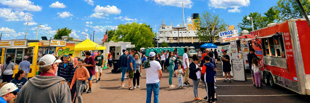 A bustling outdoor food truck festival with people walking, eating, and ordering at colorful trucks. Blue sky with scattered clouds, green trees, and distant spring waterfalls frame a large ship in the background.