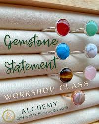 Six rings with colorful gemstone settings are displayed on a beige cushion. Text reads: "Gemstone Statement Workshop Class, Alchemy, 1204 N. 12th St. Superior, WI 54880.