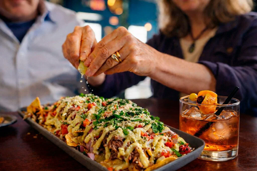 Two people sit at a table, one squeezing a lime over loaded nachos topped with cheese, tomatoes, and herbs—an inviting scene perfect for weekend getaways in Wisconsin. Nearby, a cocktail with an orange garnish completes the relaxed vibe.