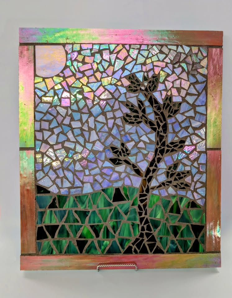 Mosaic artwork featuring a tree silhouette with brown branches, green grass, a bright sun, and a blue sky, all created with colorful, irregular glass pieces in a wooden frame.