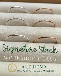 Display of five delicate stacked rings in silver and rose gold on a fabric ring holder. Text reads: “Signature Stack Workshop Class” and “Alchemy, 2724 N. 21 St. Superior, WI 54880.”.