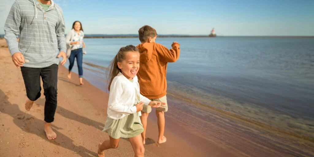A family with two children runs barefoot along a sandy beach by the water on a sunny day, smiling and enjoying their time together—capturing the joy of weekend getaways in Wisconsin.