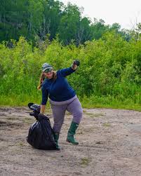 A person in outdoor clothing and boots holds a large trash bag and picks up litter in a natural area with green trees in the background.