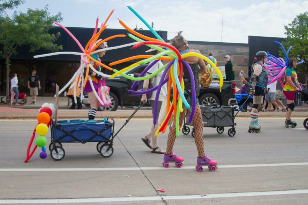 People wearing roller skates and colorful balloon wings participate in a lively parade, with wagons decorated with rainbow balloons and others walking on the street in festive, vibrant attire.