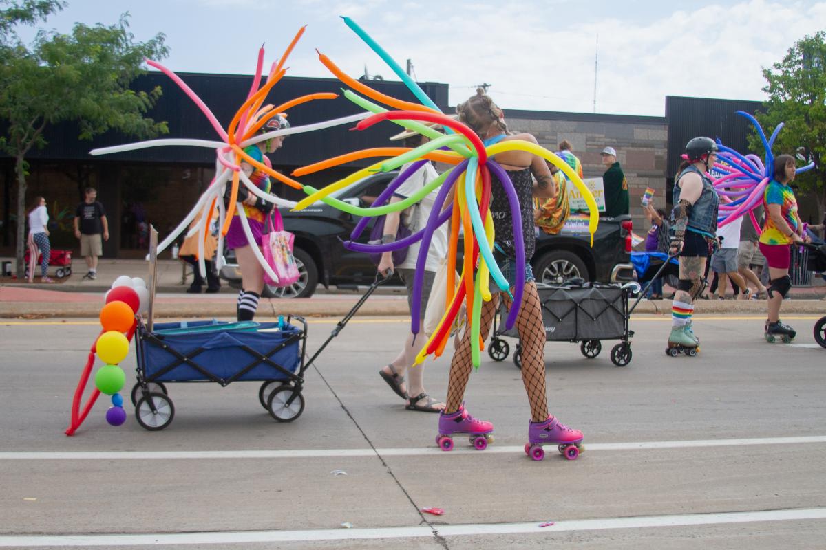 People wearing roller skates and colorful balloon wings participate in a lively parade, with wagons decorated with rainbow balloons and others walking on the street in festive, vibrant attire.