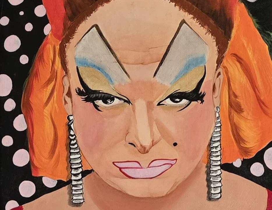 A person with dramatic stage makeup, bold angular eyebrows, blue and yellow eyeshadow, large pink lips, and orange hair is wearing large silver earrings, in front of a black background with white polka dots.