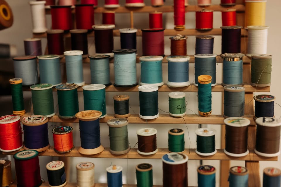 A wooden rack filled with colorful spools of thread, neatly arranged in rows. The spools vary in color, including shades of blue, green, red, brown, and beige.
