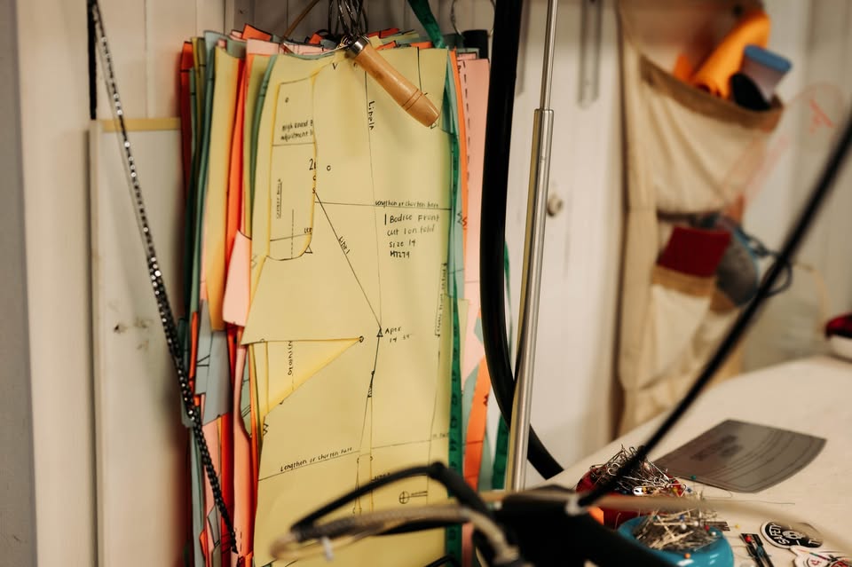 A stack of colorful sewing patterns with handwritten notes is organized on clipboards and hangs on a hook in a craft room, with various tools and supplies visible in the background.