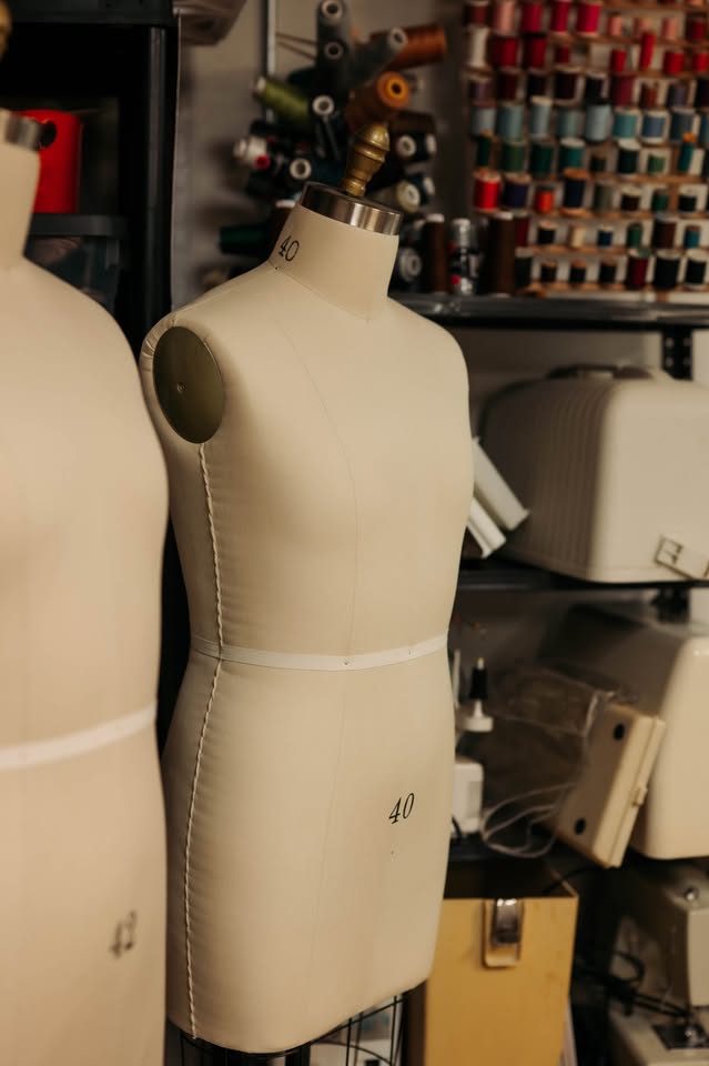 Two beige dress forms labeled with the number 40 are in a sewing studio, surrounded by shelves holding colorful spools of thread, sewing machines, and other tailoring supplies.