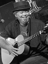 Black and white photo of a smiling man wearing a hat, seated, playing an acoustic guitar. A microphone is visible to the left, and the background includes blurred signage and lights.