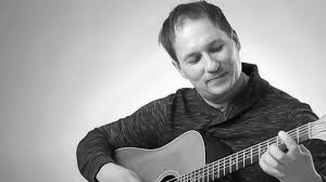 A person with short hair smiles while playing an acoustic guitar. The photo is in black and white, and the background is plain and light-colored.