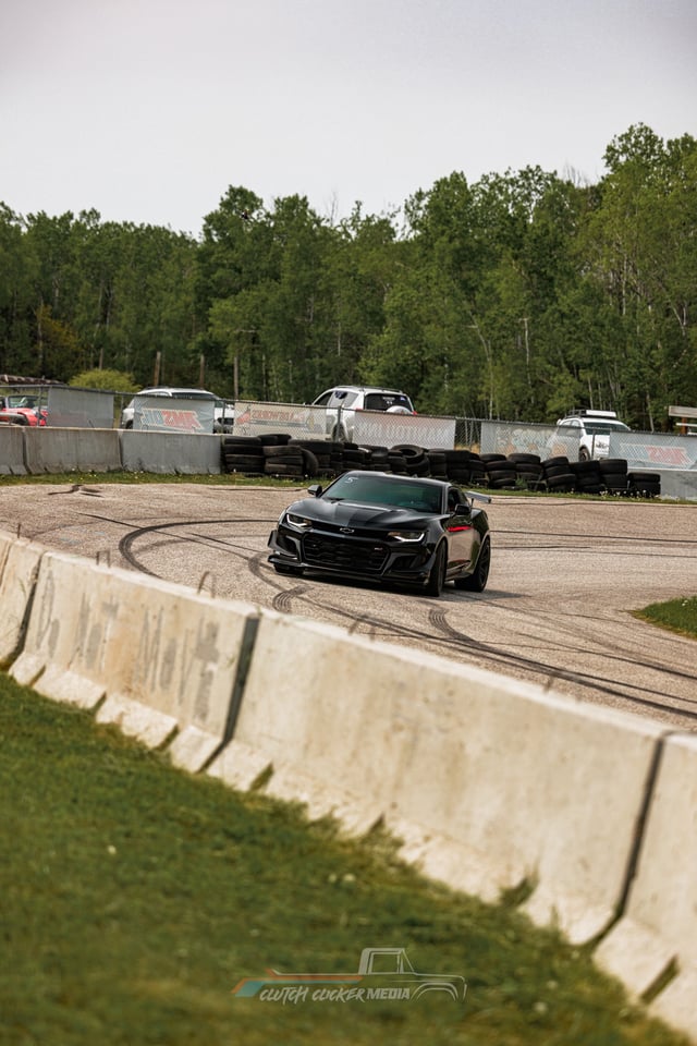 A black sports car drifts around a racetrack corner, leaving tire marks on the pavement. In the background, trees and parked trucks are visible behind a concrete barrier.