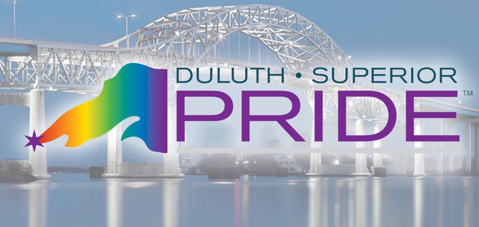 The Duluth-Superior Pride logo with a rainbow flag and star is overlaid on a photo of the illuminated Aerial Lift Bridge reflected in the water at dusk.