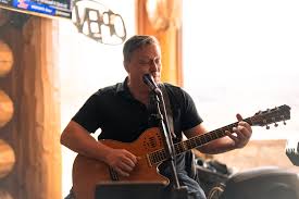A man in a black shirt plays an acoustic guitar and sings into a microphone indoors, with warm lighting and musical equipment visible in the background.