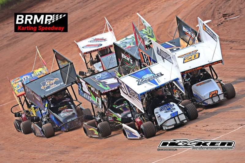 A group of go-karts with winged roofs races closely on a dirt track at BRMP Karting Speedway, with vibrant colors and sponsor logos visible on the vehicles.