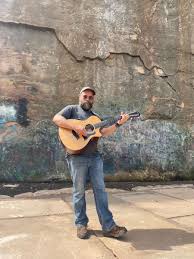 A man with a beard, wearing a cap, t-shirt, jeans, and boots, stands outdoors playing an acoustic guitar in front of a weathered, graffiti-covered concrete wall.