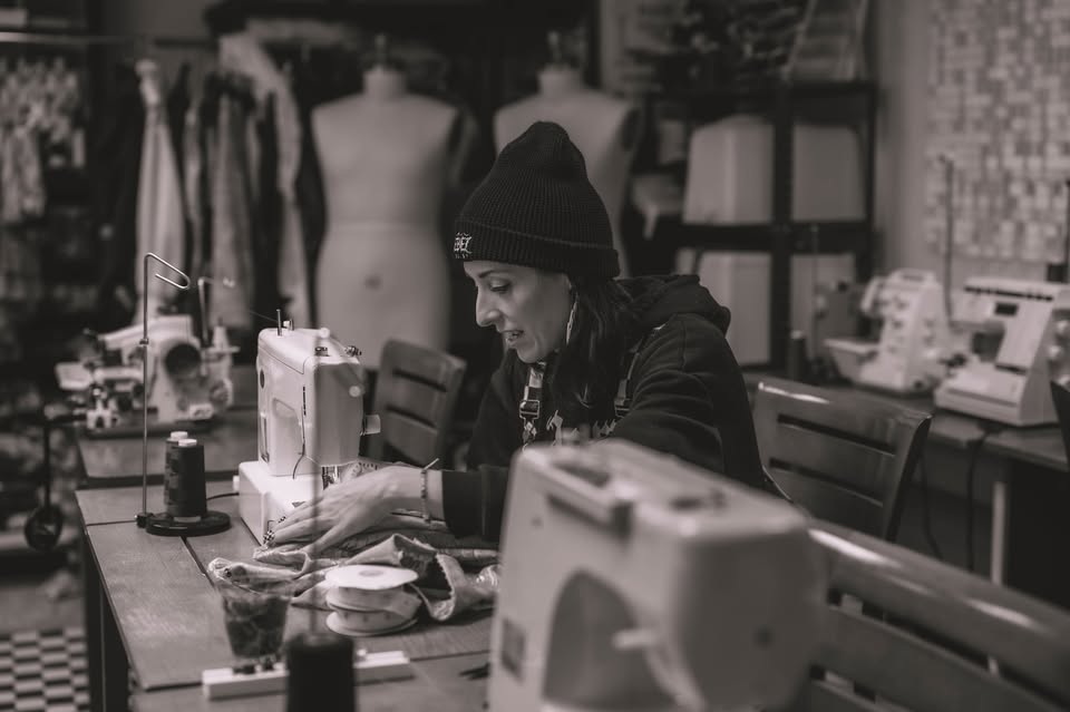 A person wearing a beanie sews fabric on a sewing machine in a workshop, surrounded by sewing equipment, thread spools, and mannequins. The image is in black and white.