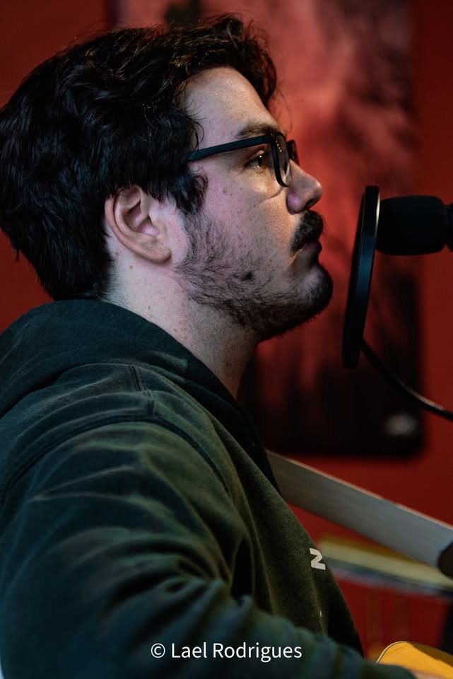 A person with dark hair, glasses, and a beard sings or speaks into a microphone. They are wearing a dark hoodie, and the background is blurred with warm colors. © Lael Rodrigues.