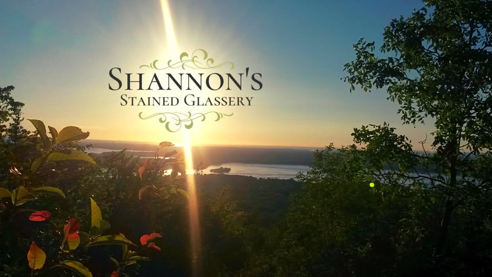 A scenic sunset view over a river and forest with "Shannon's Stained Glassery" written in decorative text in the sky. Sunlight reflects on the water and foliage frames the foreground.
